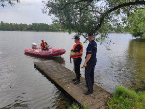 Funkcjonariusz policji wspólnie z Państwową Strażą Pożarna stoją na pomoście i obserwują jezioro.