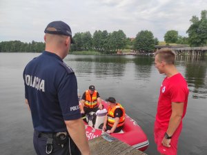 Policjant stojący na pomoście podczas wspólnych działań z ratownikiem Wodnego Ochotniczego Pogotowia Ratunkowego i Strażą Pożarną.